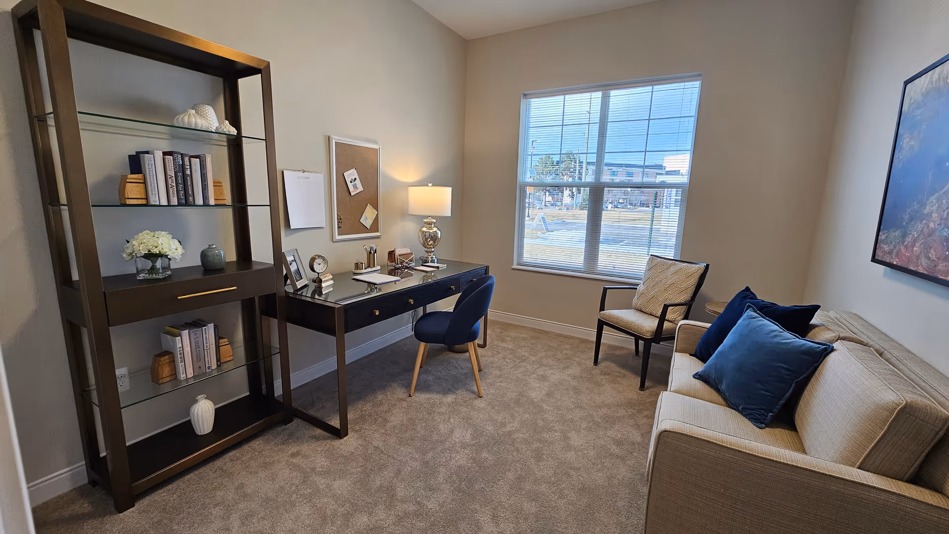A cozy interior room with a beige carpet and light-colored walls. The room features a dark wooden desk with a glass top, a blue upholstered chair, a silver table lamp, and various desk accessories. To the left is a tall shelving unit with books and decorative items. On the right side, there is a beige sofa with blue pillows and a beige armchair with a patterned cushion. A large window with blinds lets in natural light, and a framed artwork hangs on the wall above the sofa.