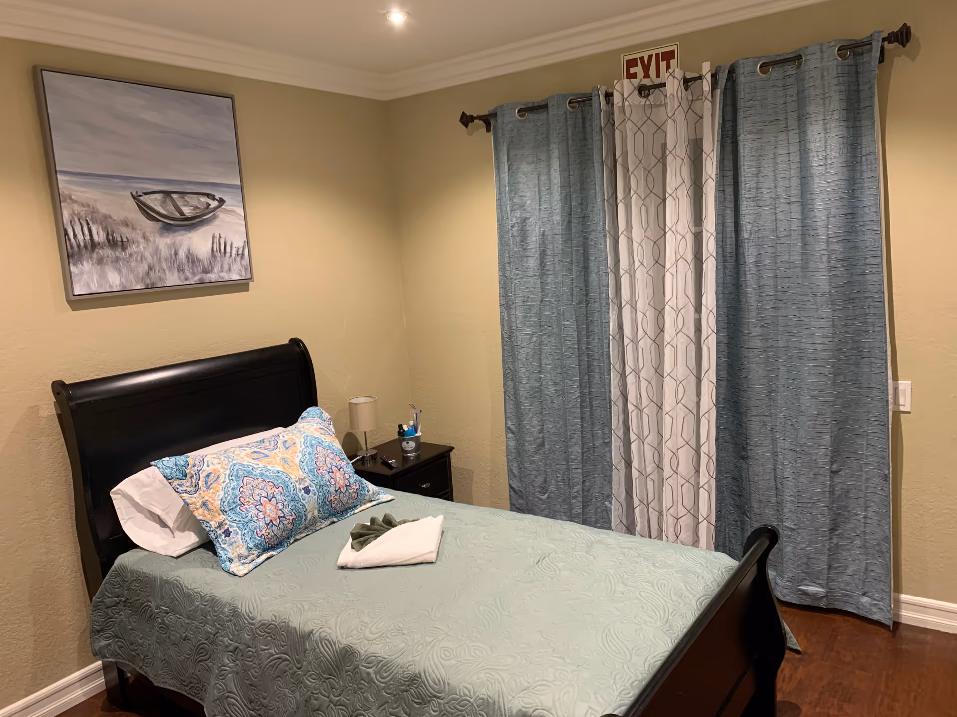 A neatly made single bed with a light green quilt and two decorative pillows in blue and yellow patterns. Next to the bed is a small dark wooden nightstand with a lamp and some small items. Behind the bed is a wall with a painting of a boat on a beach. A window with blue and white curtains is visible, with an exit sign above it. The room has beige walls and wooden flooring.