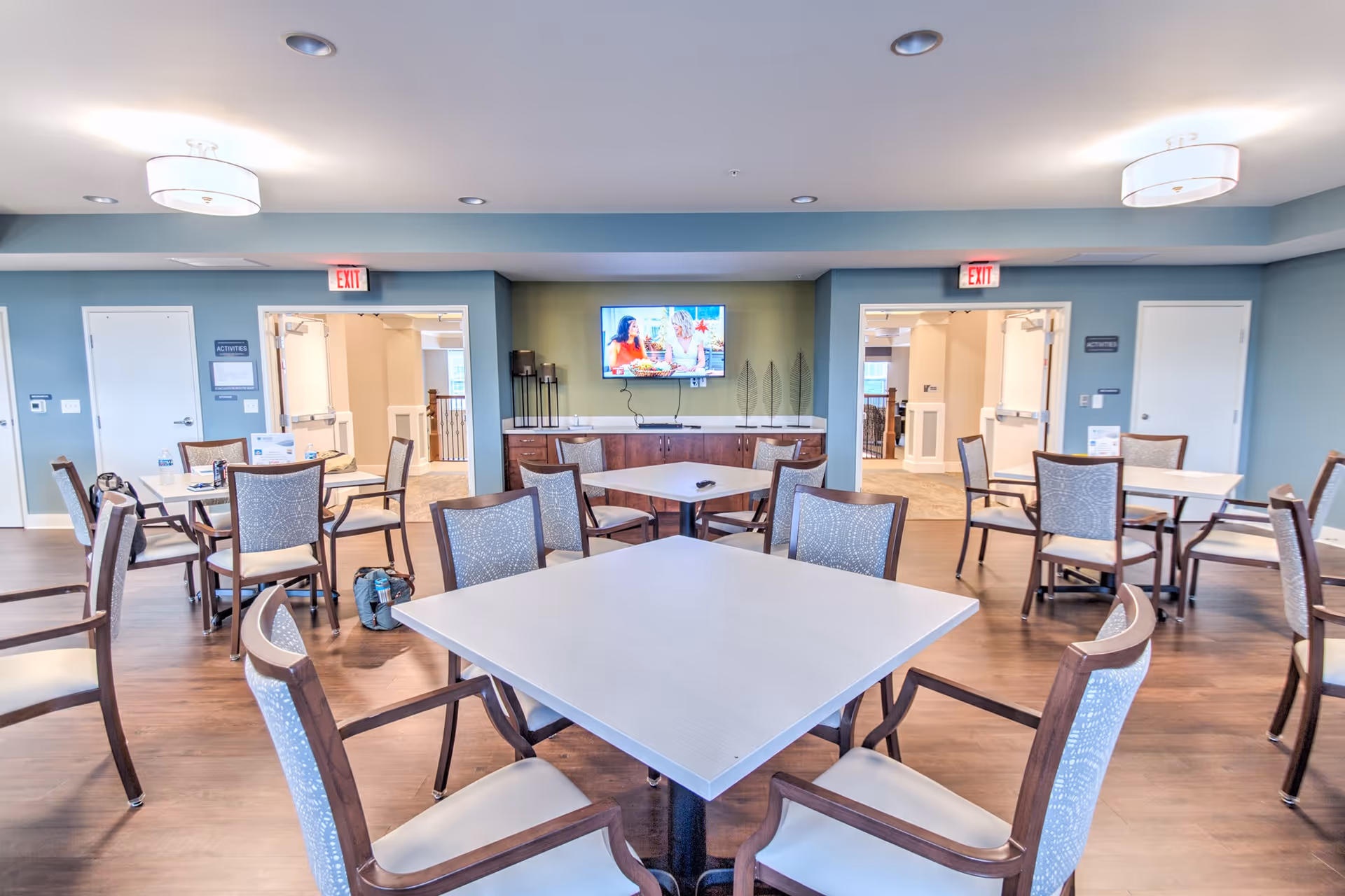 A bright and spacious common area in a senior living facility with multiple square tables and cushioned chairs arranged around the room. A flat-screen TV is mounted on a green accent wall above a wooden cabinet. The walls are painted blue and white, and there are exit signs above two doorways leading to other parts of the building. The floor is wood, and the ceiling has modern light fixtures.
