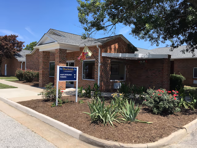 Exterior view of a single-story brick building with a sign that reads 'Dockside Health & Rehab Center' indicating the main entrance and parking. The building is surrounded by landscaped flower beds and a sidewalk under a clear blue sky.