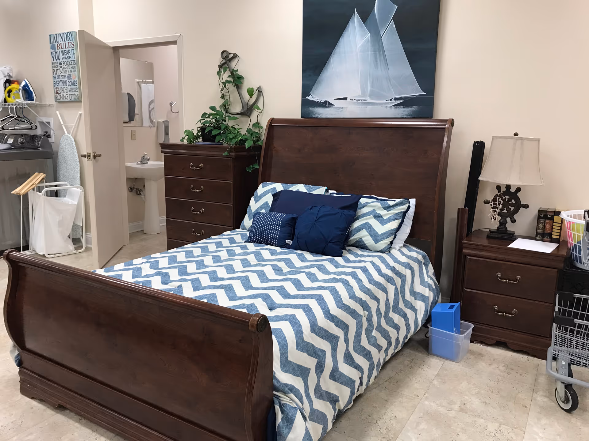A bedroom with a wooden bed frame and a blue and white chevron patterned bedspread. There are several blue pillows on the bed. To the right of the bed is a wooden nightstand with a lamp, books, and a basket. To the left is a wooden dresser with a plant on top. A painting of a white sailboat on a dark background hangs above the bed. In the background, there is an open door leading to a small bathroom with a sink and mirror. Laundry supplies and an ironing board are visible near the bathroom door.