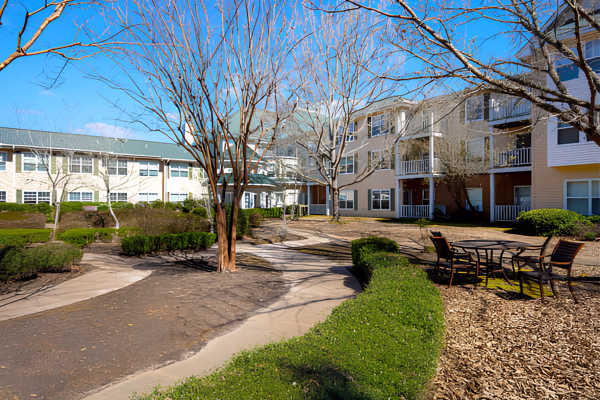 Courtyard with leafless trees, paved walkways and a patio table set in front of a multi-story assisted living building under a clear blue sky.