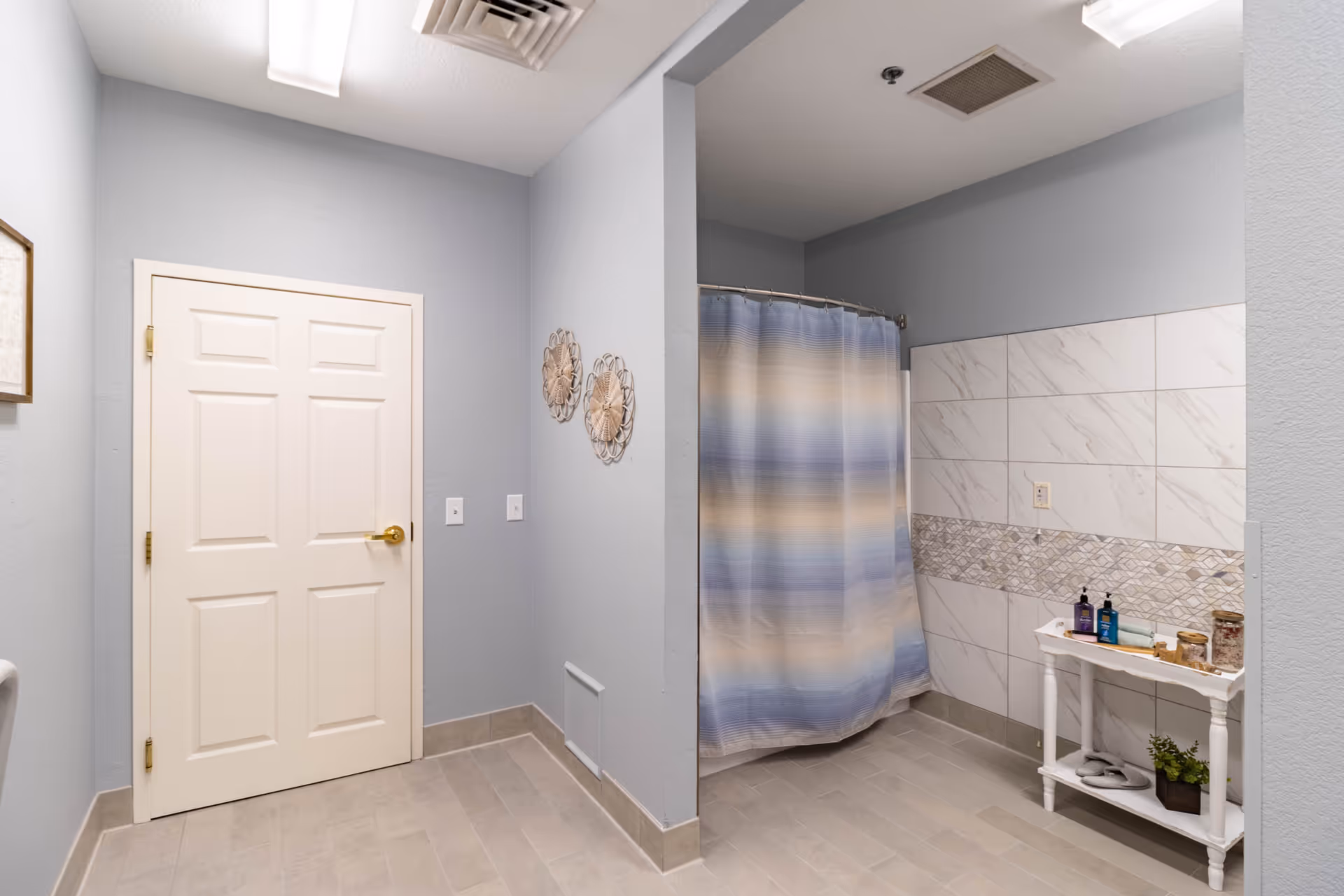 A clean bathroom with light blue walls and beige tiled floor. There is a white door with a gold handle on the left side, and a shower area with a blue and beige striped shower curtain on the right. The shower area has white and beige tiled walls with a decorative tile strip. A small white shelf holds toiletries and decorative items near the shower.