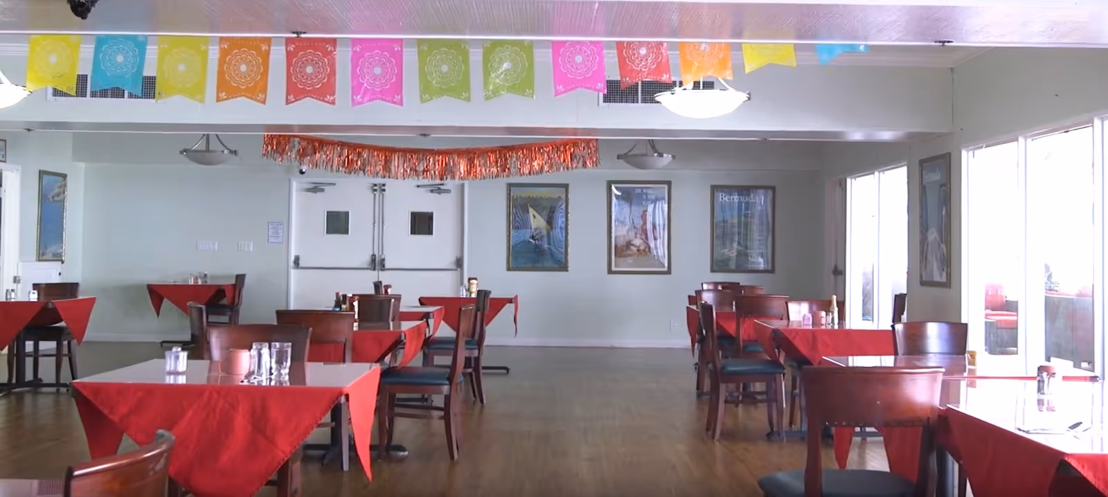 A dining room with several wooden tables and chairs, each table covered with a red tablecloth. The room has large windows on the right side letting in natural light. Colorful decorative banners hang from the ceiling, and framed pictures are on the far wall. The floor is wooden, and the room appears clean and well-lit.