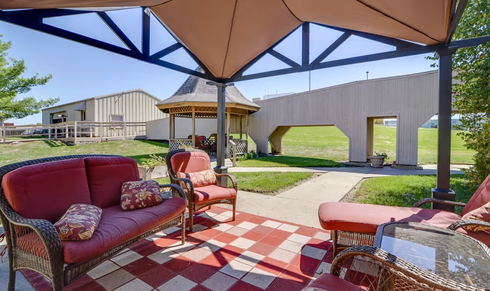 Patio with red cushioned wicker seating under a canopy, a gazebo, and a grassy lawn with nearby buildings.