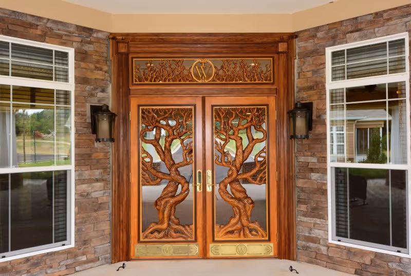 Ornate wooden double doors with intricate tree carvings, flanked by two windows and mounted wall lanterns, set in a stone exterior wall.
