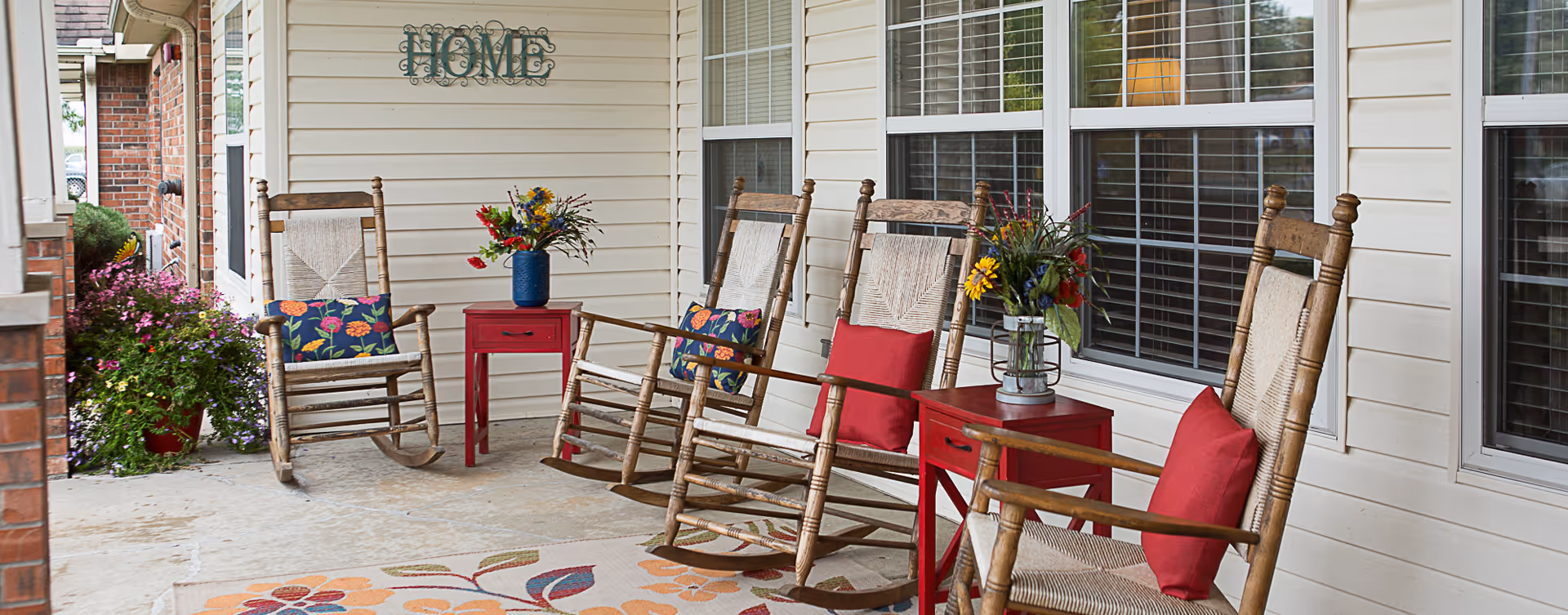 A covered outdoor porch area with four wooden rocking chairs, each with a red or floral cushion. Two small red side tables hold vases with colorful flowers. A decorative sign that says 'HOME' is mounted on the white siding wall. There are windows with blinds behind the chairs and a floral rug on the floor.