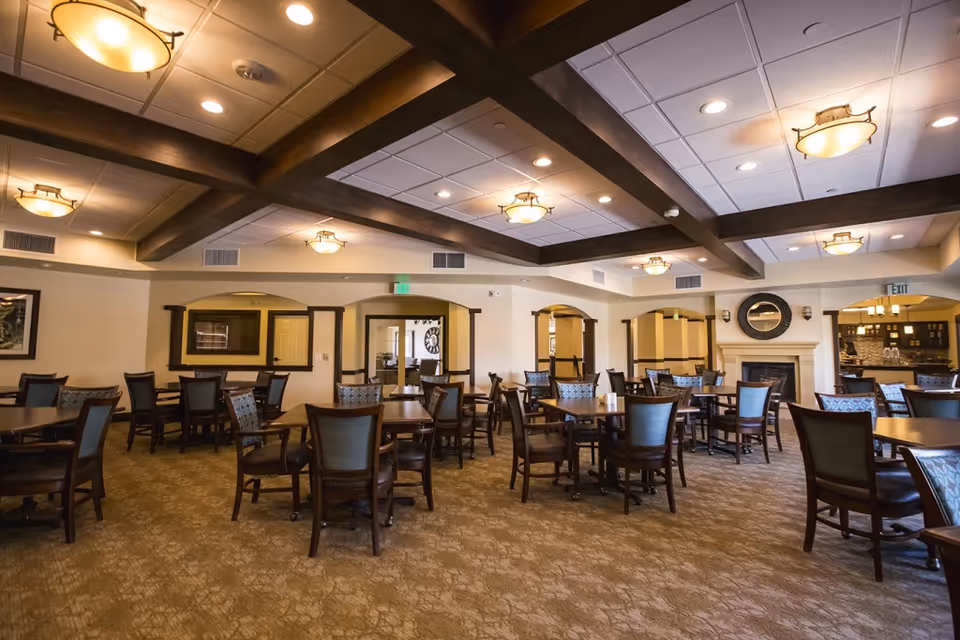 Spacious senior living dining room with multiple wooden tables and chairs, exposed ceiling beams, warm lighting, and a fireplace.