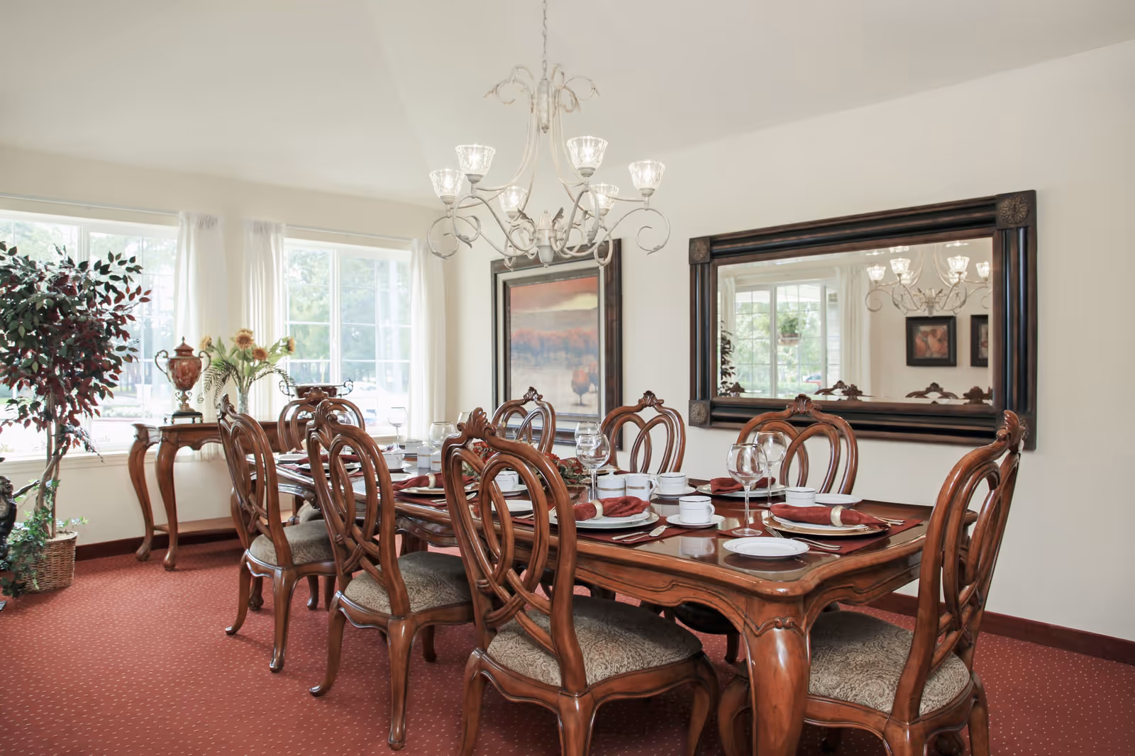 A formal dining room with a large wooden dining table set for eight people. The table is adorned with white plates, wine glasses, and red napkins. The room has a red carpet, a large mirror on the wall, a chandelier overhead, and a window with white curtains letting in natural light. There is also a small wooden side table with decorative items and a potted plant in the corner.