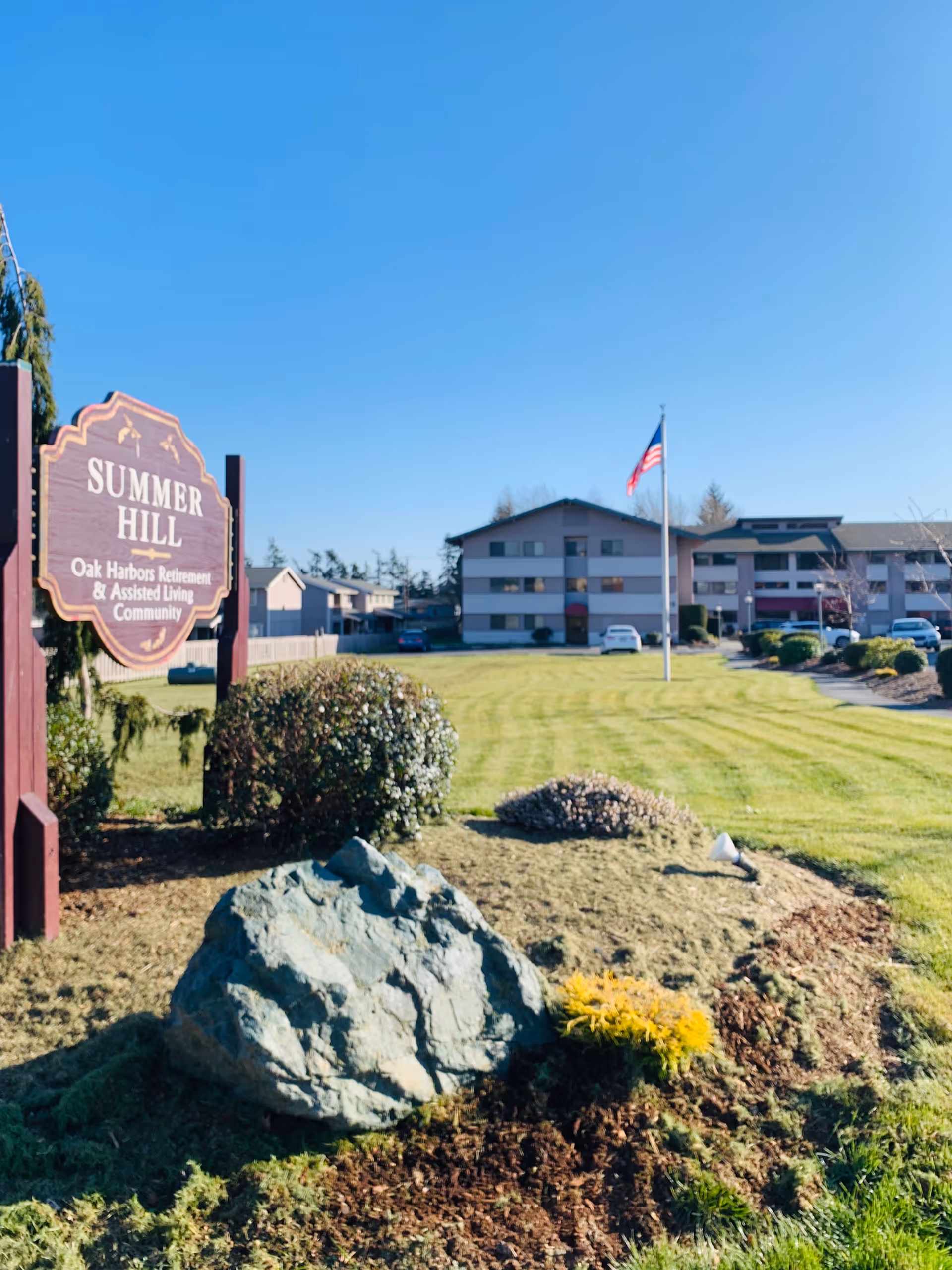 Outdoor view of Summer Hill Oak Harbors Retirement & Assisted Living Community with a large sign in the foreground, a rock and landscaped bushes, a green lawn, an American flag on a flagpole, and a multi-story building in the background under a clear blue sky.
