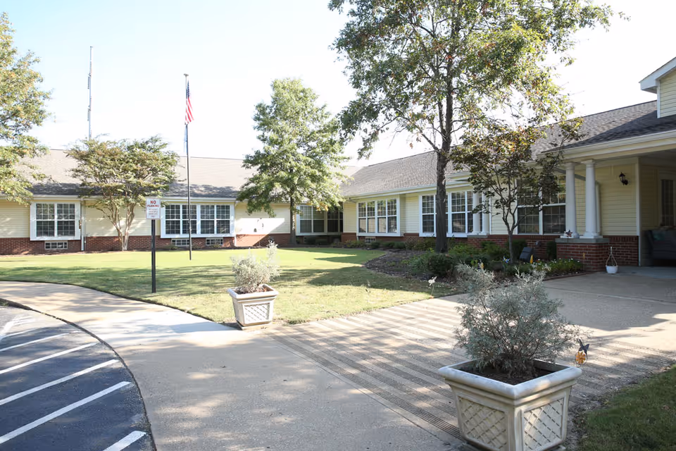 Exterior view of Regency Retirement Village of Jackson showing a single-story building with multiple windows, a covered entrance supported by white columns, a well-maintained lawn with trees and shrubs, two large planters with plants, and a flagpole with an American flag.