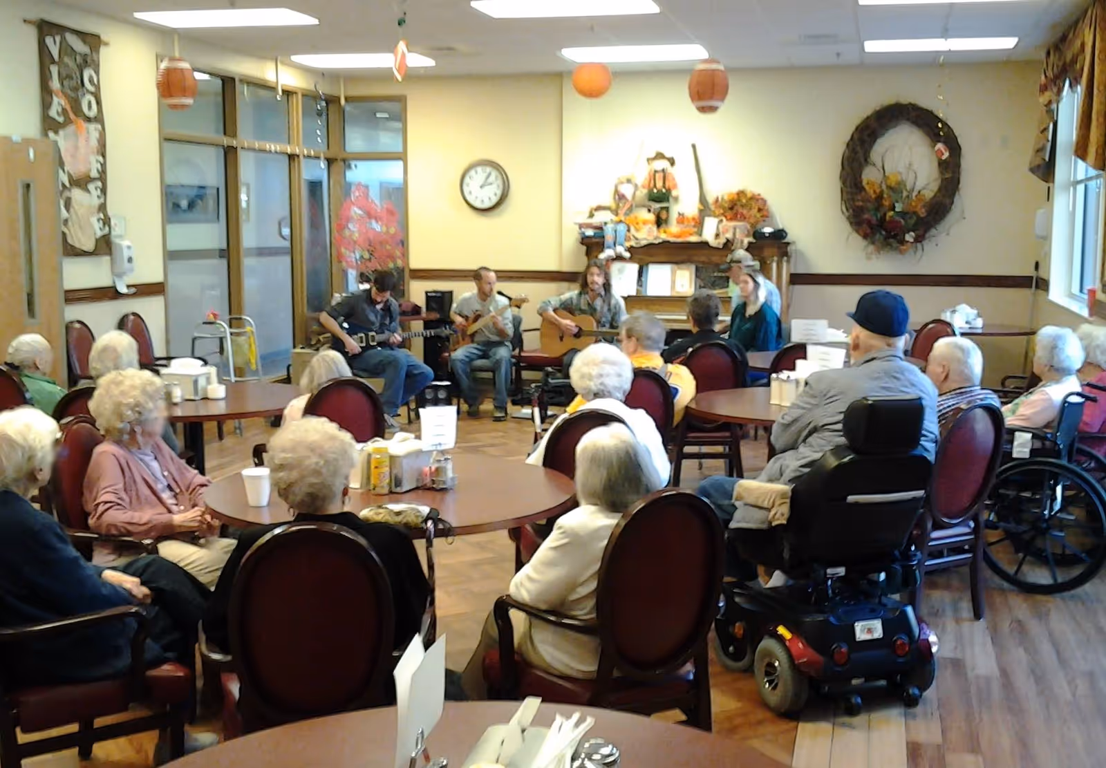 A group of elderly people seated around tables in a common room, watching a small band of musicians playing guitars and singing. The room is decorated with autumn-themed items and a clock on the wall shows the time. Some residents are in wheelchairs and one is in a motorized scooter.