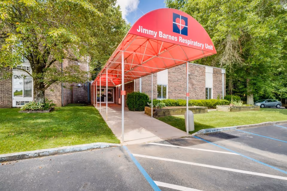Entrance to the Jimmy Barnes Respiratory Unit at Greystone Healthcare Center, featuring a red canopy over a sidewalk leading to the building entrance, surrounded by green grass, trees, and a parking area with marked spaces.