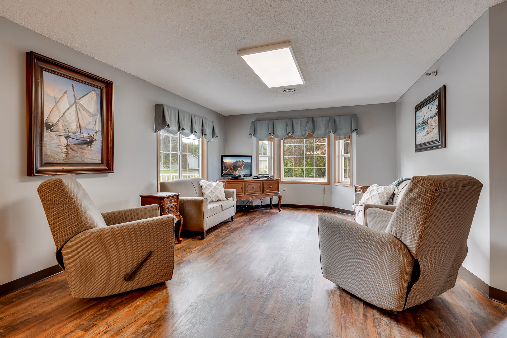 A cozy living room with two beige armchairs and two beige sofas arranged around a wooden floor. The room has large windows with blue valances letting in natural light. There are two framed paintings on the walls and a wooden cabinet with a TV on top.