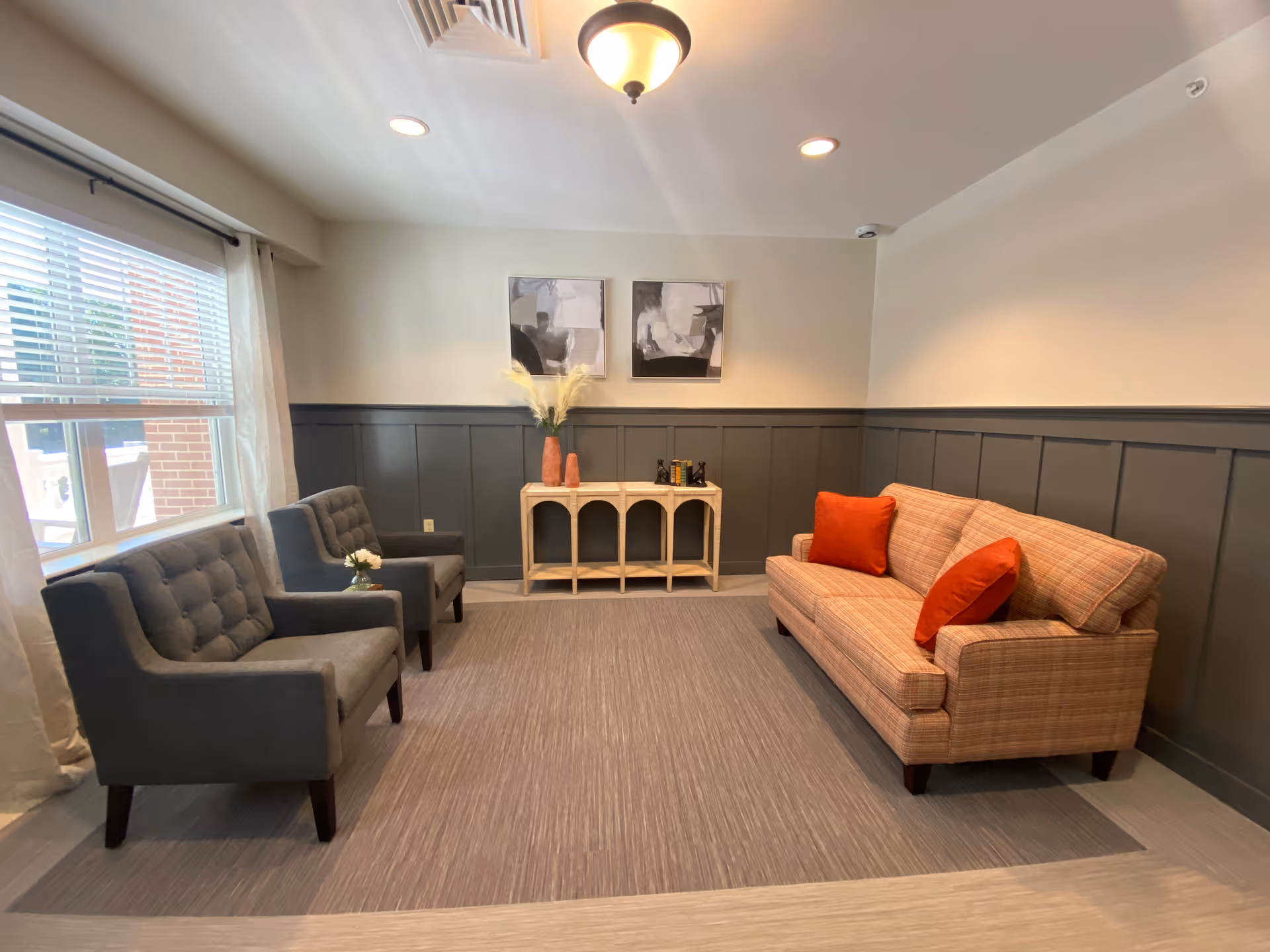 A cozy sitting area with a plaid sofa adorned with two orange pillows on the right, and two gray tufted armchairs on the left. Between the chairs and sofa is a light-colored console table with decorative vases and books. The walls are painted white on top and dark gray on the bottom with paneling. Two abstract paintings hang above the console table. A window with white curtains lets in natural light from the left side.