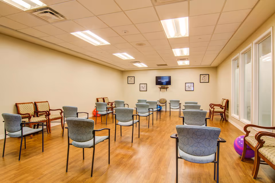A spacious room with wooden flooring and beige walls, arranged with multiple chairs spaced apart in rows facing a wall-mounted television. The room has fluorescent ceiling lights, framed pictures on the walls, and some exercise balls placed near the walls. There are also windows on the right side of the room.