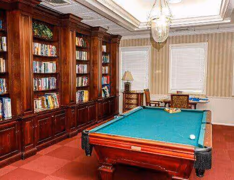 A room with a green felt pool table in the center, surrounded by wooden bookshelves filled with books along one wall. The room has two windows with blinds, a table with chairs near the windows, and a ceiling light fixture above the pool table.