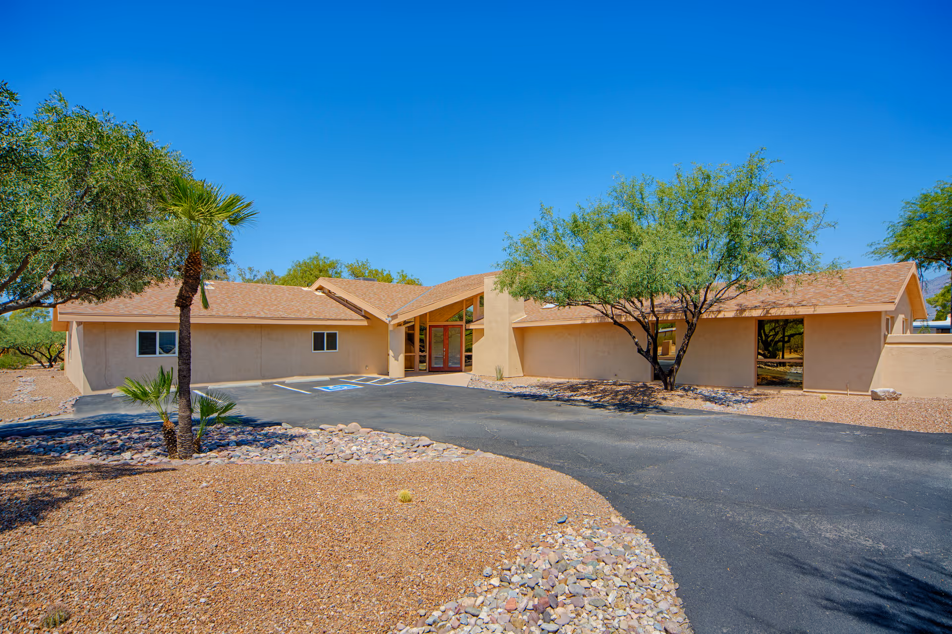 Exterior view of a single-story beige building with a sloped roof, surrounded by desert landscaping with rocks, small palm trees, and other desert plants under a clear blue sky.