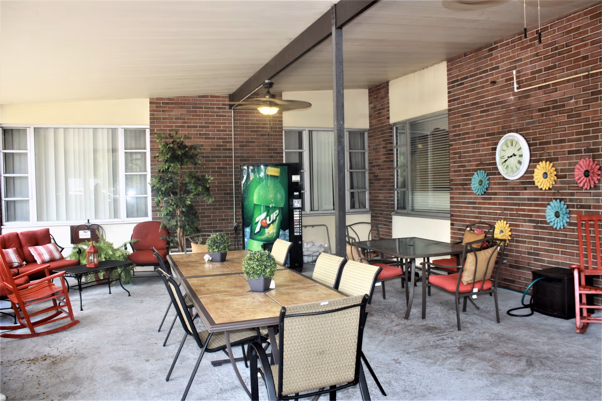 Covered outdoor patio area with multiple tables and chairs, including cushioned seating and rocking chairs. A 7UP vending machine is visible against a brick wall decorated with colorful flower-shaped wall hangings and a clock. There are potted plants on the tables and a ceiling fan overhead.