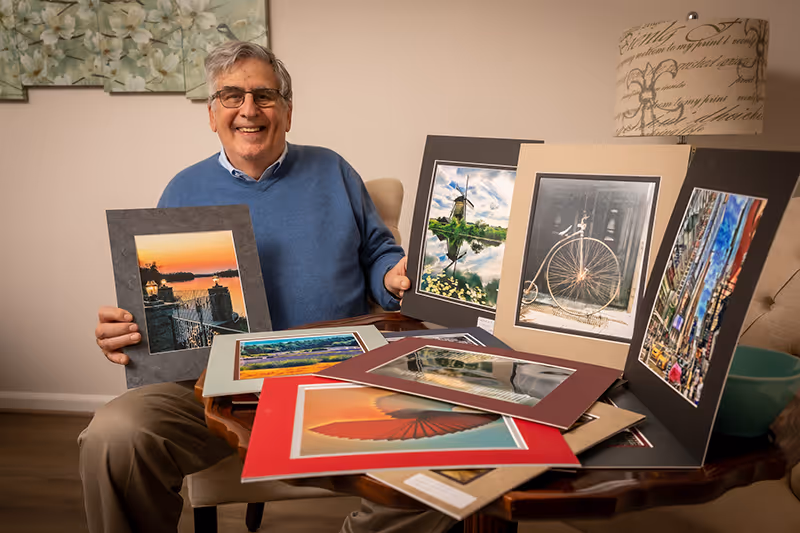 An elderly man wearing glasses and a blue sweater sits at a wooden table displaying several framed photographs and artwork. He is smiling and holding one of the framed pictures. The room has light-colored walls, a floral painting, and a decorative lamp in the background.