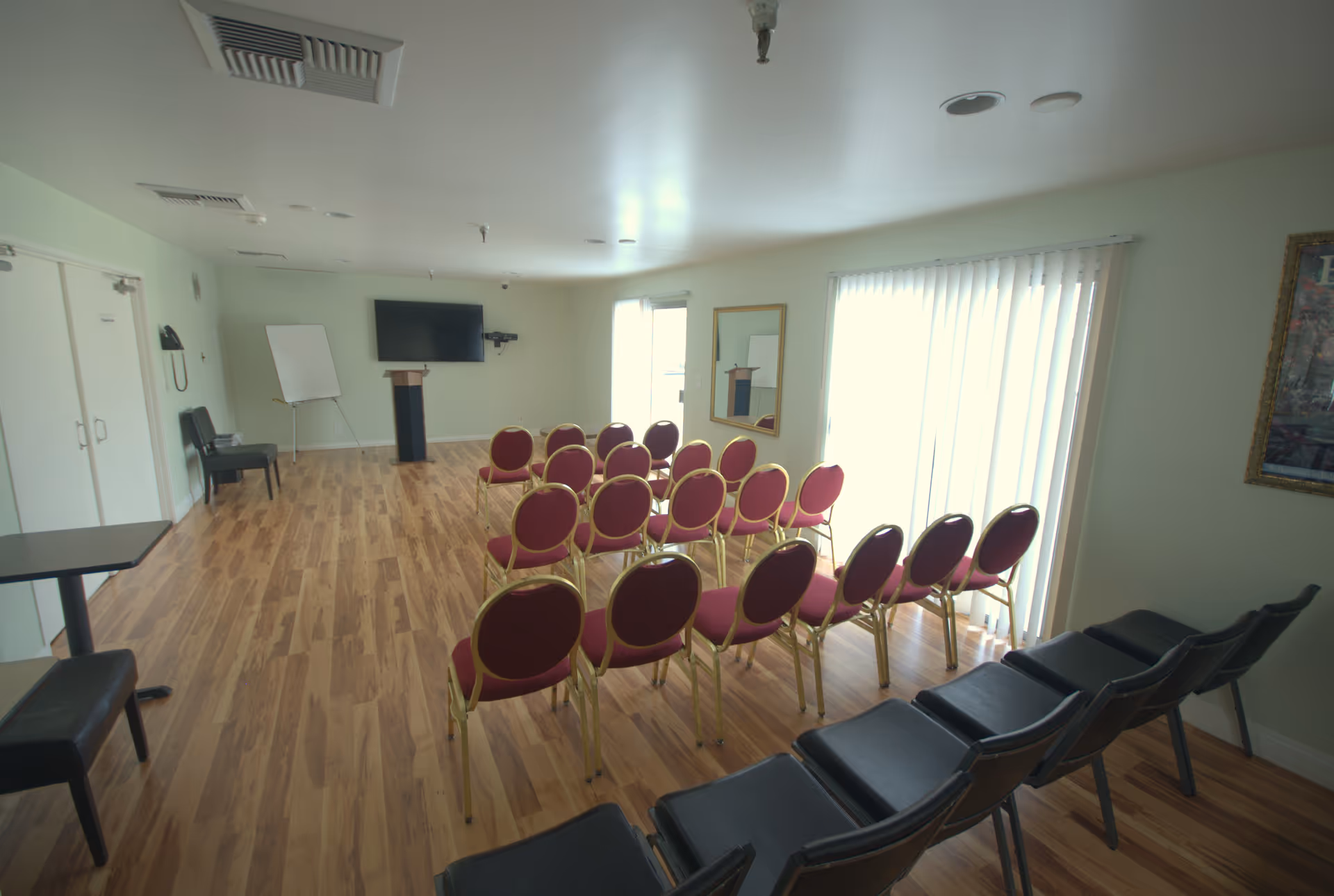 A meeting or presentation room with rows of red and black chairs facing a podium and a wall-mounted TV screen. The room has wooden flooring, light green walls, a whiteboard on a stand, and vertical blinds covering a large window or glass door.