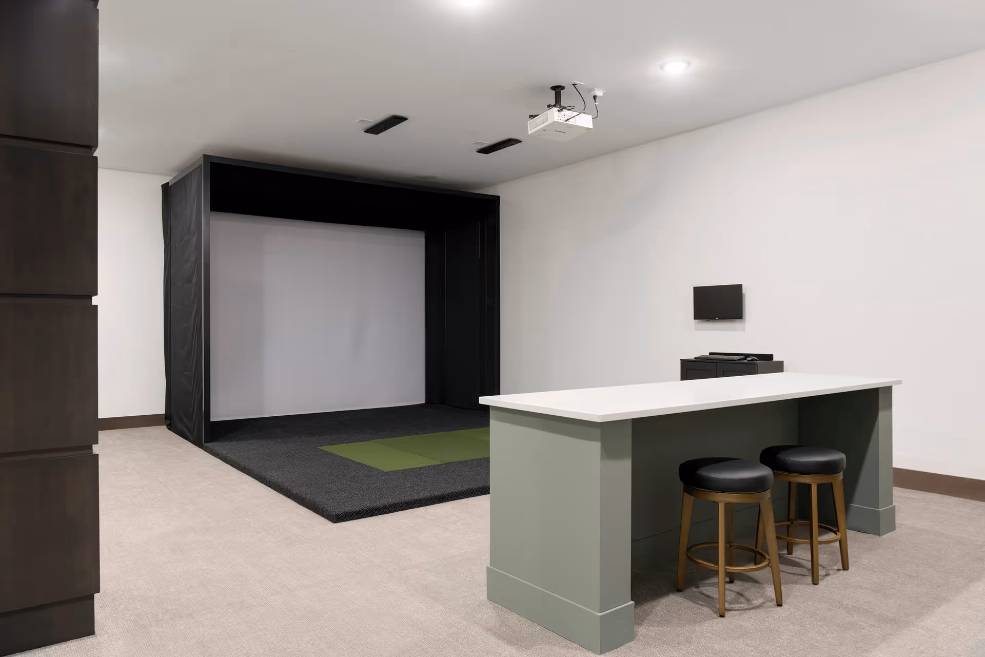 Minimal recreational interior with a projector and large screen simulator bay, a long island counter with two stools, and a small wall-mounted monitor.