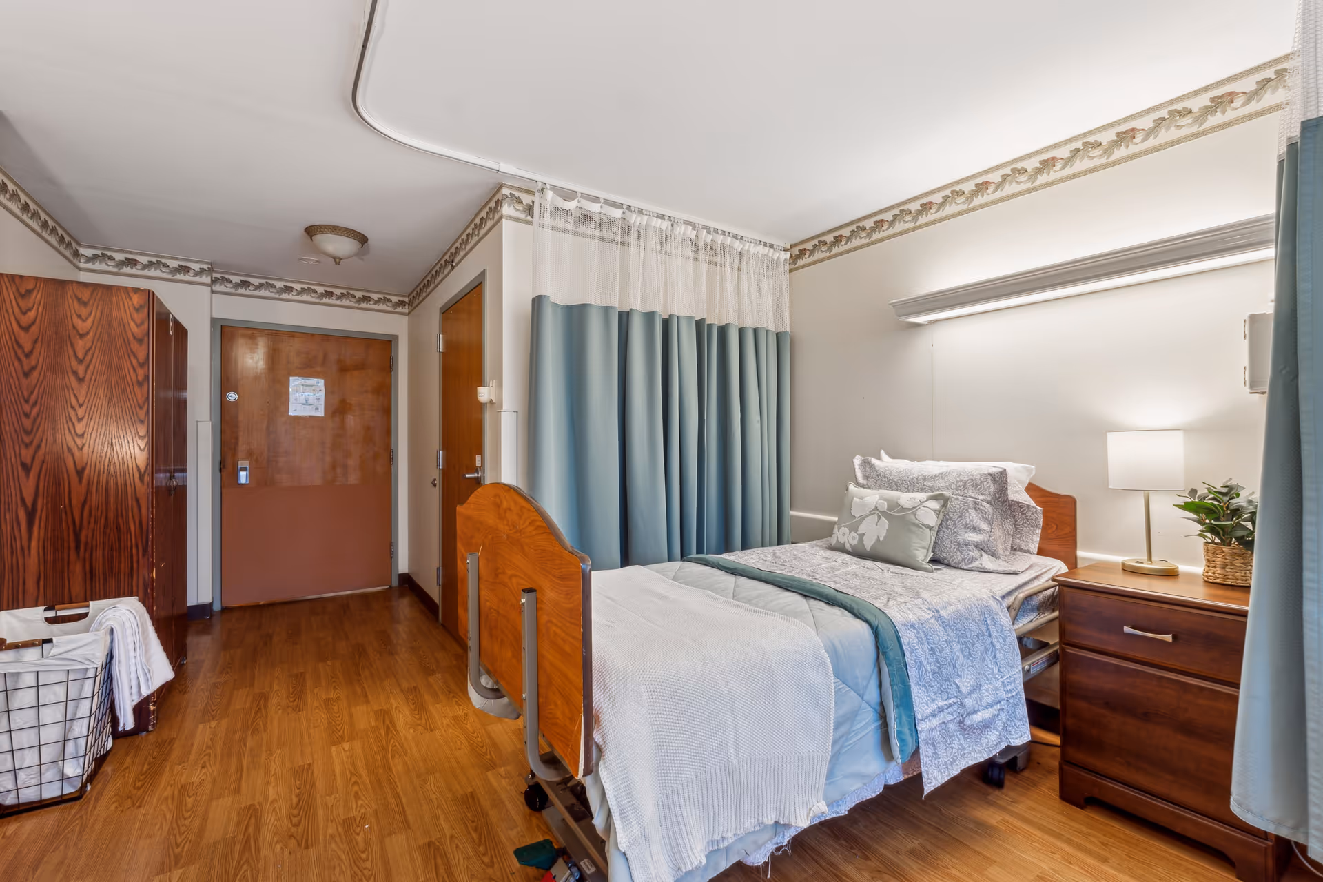 A nursing facility bedroom with a single bed made with white and blue bedding, a wooden headboard and footboard, and a bedside table with a lamp and a small plant. The room has wood flooring, a wooden wardrobe, a laundry basket with towels, and a door at the far end. A blue privacy curtain is partially drawn around the bed.