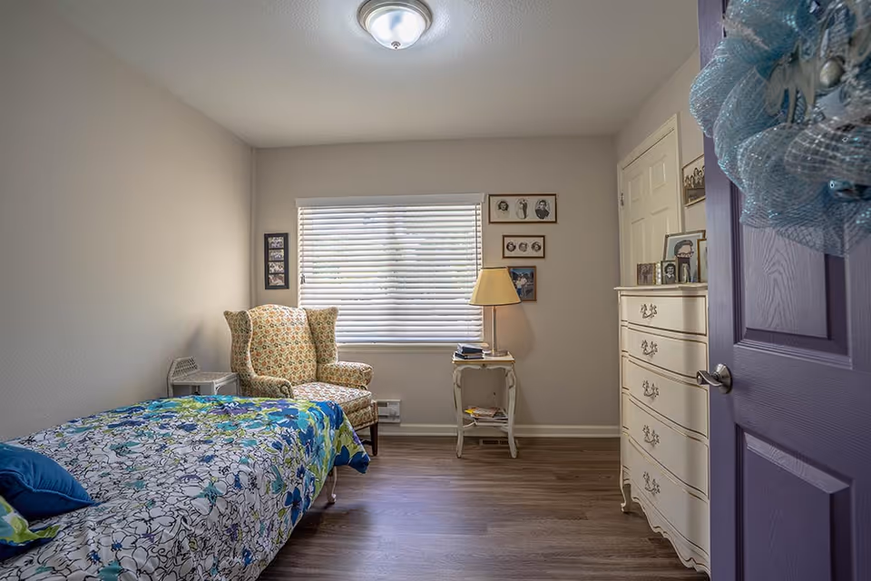 A cozy bedroom with a single bed covered in a floral bedspread, a cushioned armchair with a floral pattern, a small white side table with a lamp, and a white dresser. The room has a window with blinds, framed pictures on the walls, and a purple door with a decorative wreath.