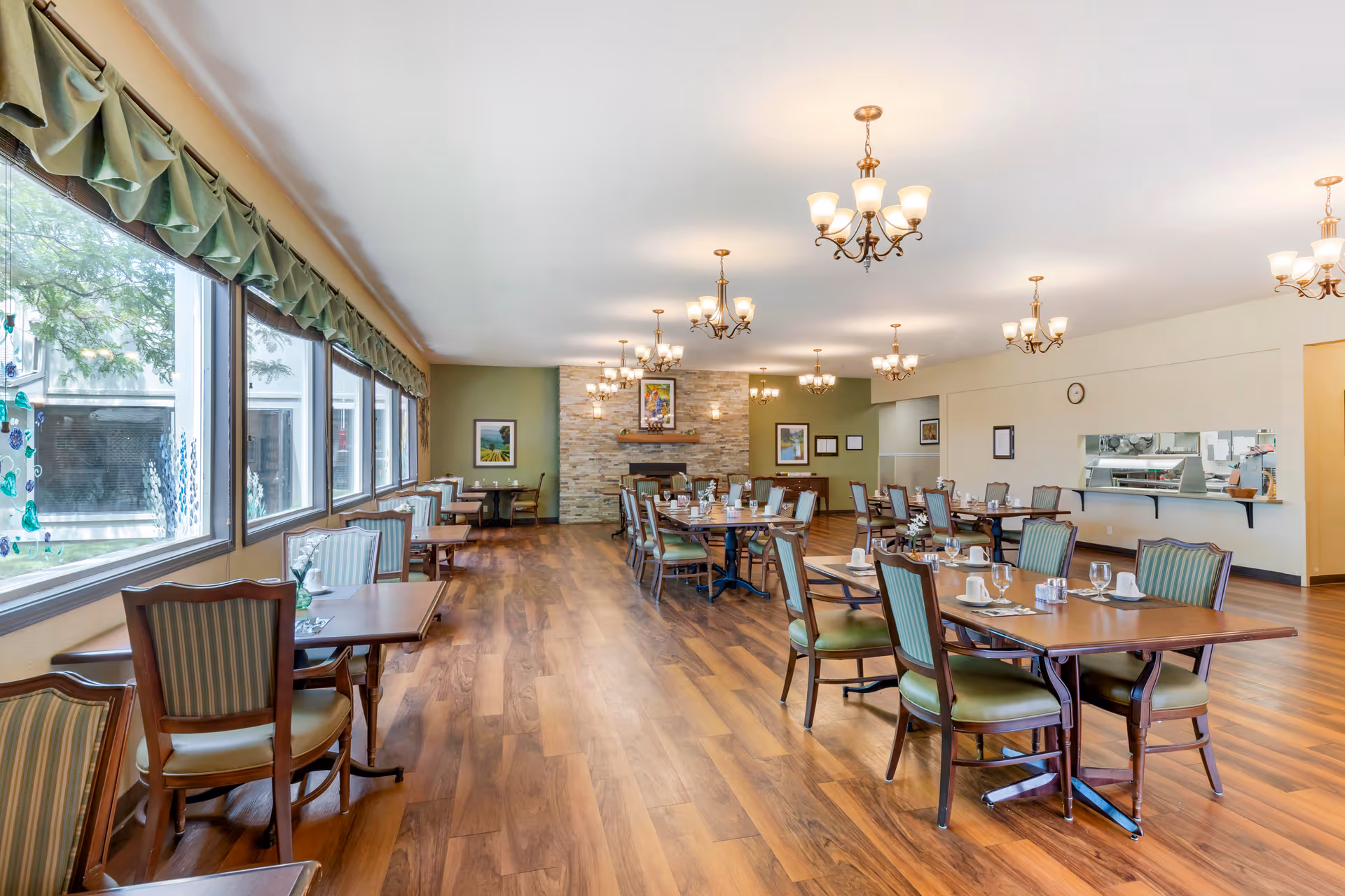 A spacious dining room with wooden floors and multiple tables set with cups, glasses, and napkins. The room has large windows on one side with green valances, several chandeliers hanging from the ceiling, and a stone fireplace at the far end with framed artwork above it. There is a serving window to the kitchen on the right wall.