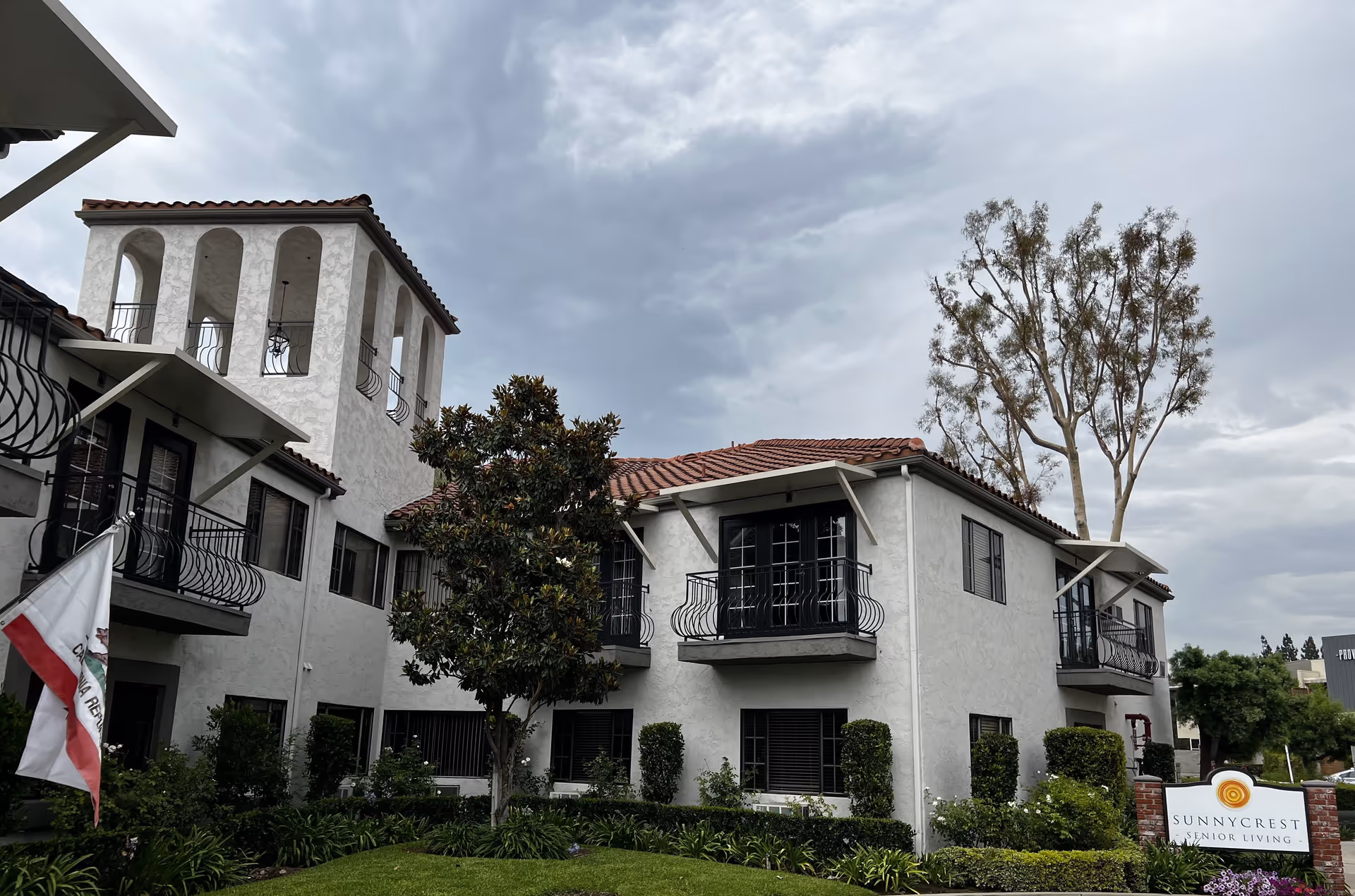 Exterior view of a two-story senior living facility building with white stucco walls and red tile roof under a cloudy sky. The building features balconies with black railings, a tower with arched openings, and surrounding greenery including trees and bushes. A sign in front reads 'Sunnycrest Senior Living'.
