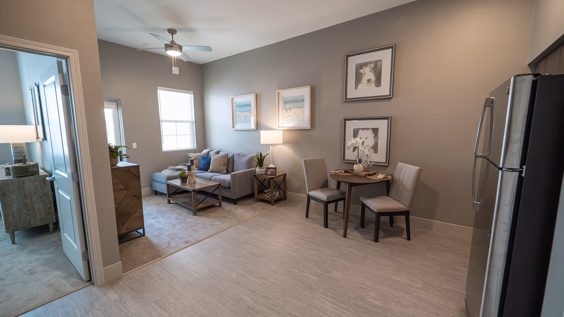 Interior view of a senior living facility showing a small dining area with a round table and two chairs, a living room with a gray sofa, coffee table, side table with a lamp, and framed artwork on the walls. A ceiling fan is visible above, and a stainless steel refrigerator is on the right side. A doorway leads to a bedroom with a dresser and lamp.