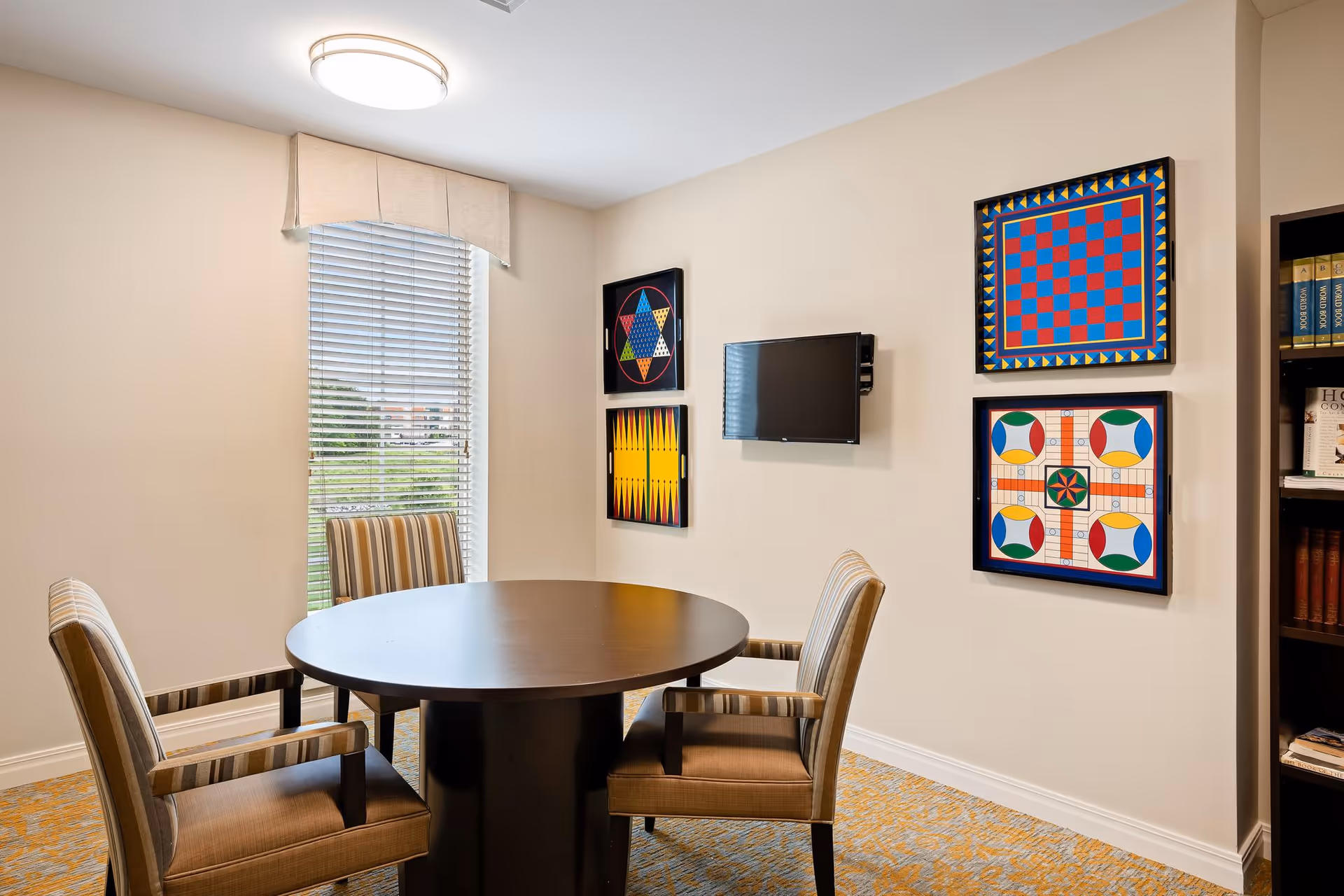 A small room with a round wooden table and four striped upholstered chairs. The walls are beige with four colorful framed game boards hanging, including checkers and other board games. A small flat-screen TV is mounted on the wall. A window with blinds and a beige valance lets in natural light. A bookshelf with books is partially visible on the right side.