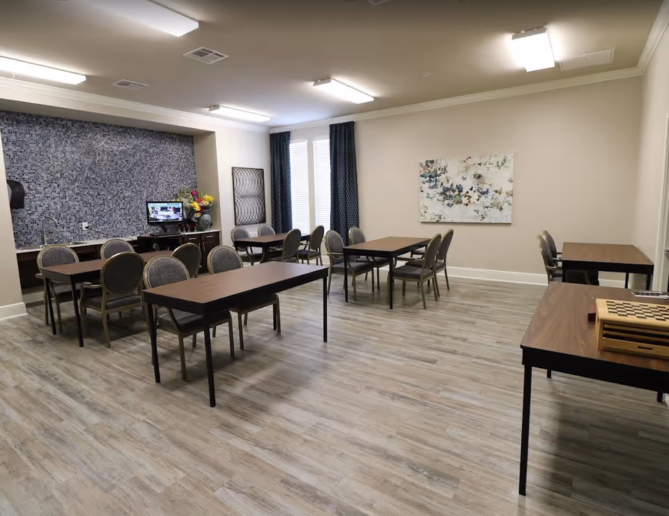 A spacious room with multiple rectangular tables and chairs arranged for group seating. The room has light-colored walls, wood-patterned flooring, and a blue mosaic tiled area with a sink and a small TV on a cabinet. There are two large windows with dark curtains and a colorful abstract painting on one wall. A chessboard is placed on one of the tables.