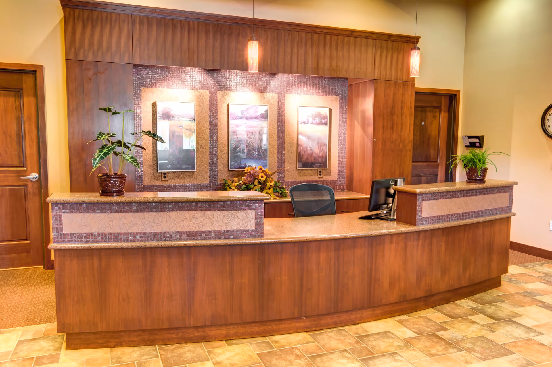 Reception desk area in a memory care residence with a wooden counter, two potted plants, a computer monitor, and three framed landscape pictures on the wall behind the desk. The floor has a tiled pattern and there are two wooden doors visible in the background.