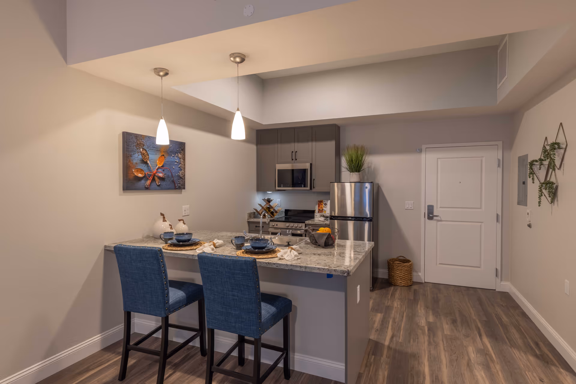 Modern kitchen area with a granite countertop island featuring two blue upholstered bar stools set with blue dishes and napkins. The kitchen includes gray cabinets, a stainless steel refrigerator, microwave, and stove. Two pendant lights hang above the island, and there is a decorative wall art and small plants on the walls. The floor is wood-style laminate, and a white door is visible in the background.
