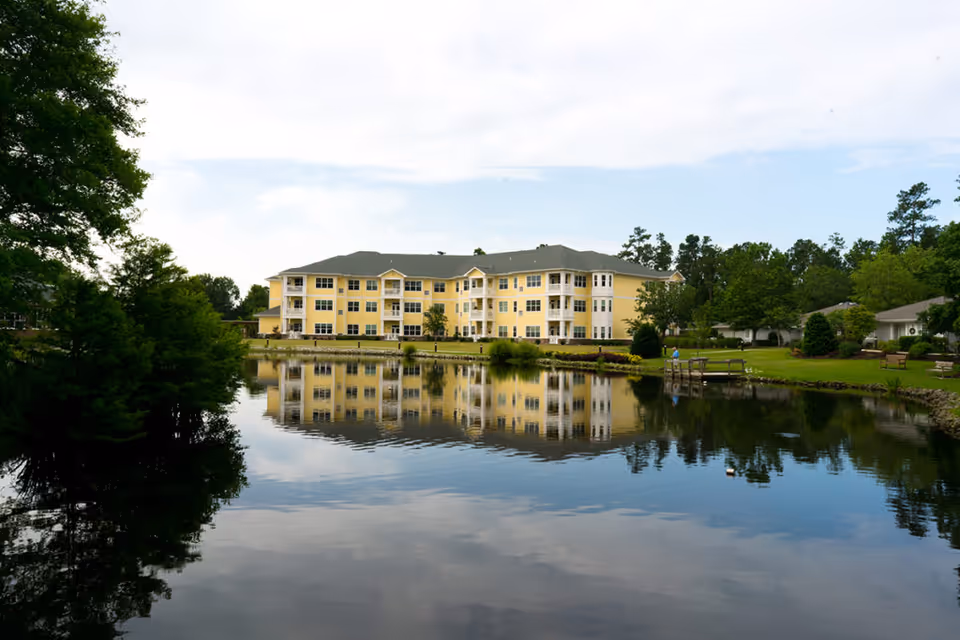 A large yellow three-story building with multiple windows and balconies reflected in a calm pond, surrounded by green trees and grassy areas with benches and a small dock.