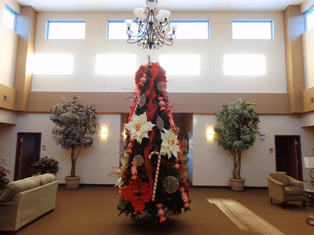 A decorated Christmas tree with red ribbons, white poinsettias, candy canes, and ornaments stands in the center of a spacious living room with high windows near the ceiling. The room has beige walls, two large potted plants on either side of the tree, a chandelier hanging above, and beige sofas and chairs arranged around the space.