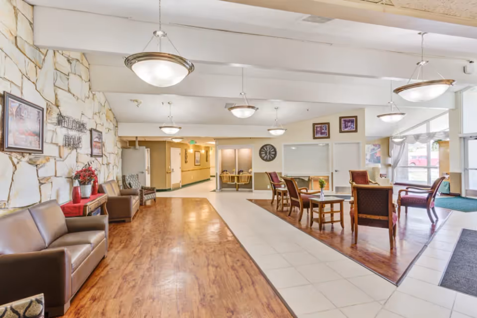 A spacious senior living facility common area with a mix of wooden and tiled flooring. The left side features a stone accent wall with framed artwork and a 'Welcome Home' sign above a small table with red vases and flowers. There are several brown leather sofas and chairs along the wall. The right side has a seating area with wooden chairs and small tables, decorated with flowers. Large windows allow natural light to fill the space, and multiple hanging light fixtures illuminate the room. The entrance door and hallway are visible in the background.