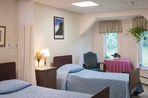 Two twin beds in a pastel senior living room with a nightstand, lamp, and window.
