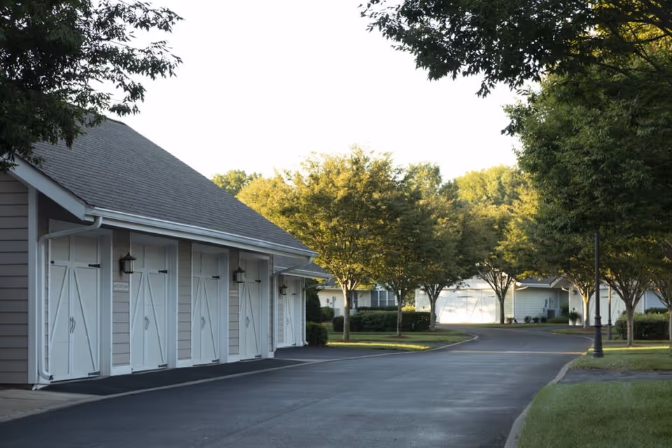 A paved driveway lined with trees and grass leads to a row of garages with white doors on the left side. Additional buildings and garages are visible in the background, surrounded by greenery under a clear sky.