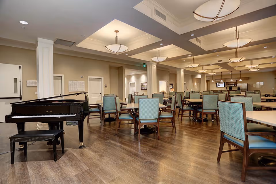 A spacious dining room with multiple tables and chairs arranged neatly. The room features a polished wooden floor, beige walls, and ceiling lights with modern fixtures. A black grand piano with a matching bench is positioned on the left side of the room.