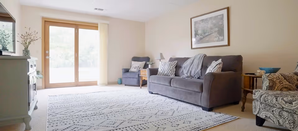 A bright living room with a large patterned rug on the floor, a gray sofa with decorative pillows and a throw blanket, a matching armchair, a patterned armchair, a wooden side table with a lamp, and a framed picture on the beige wall. There are glass sliding doors letting in natural light.