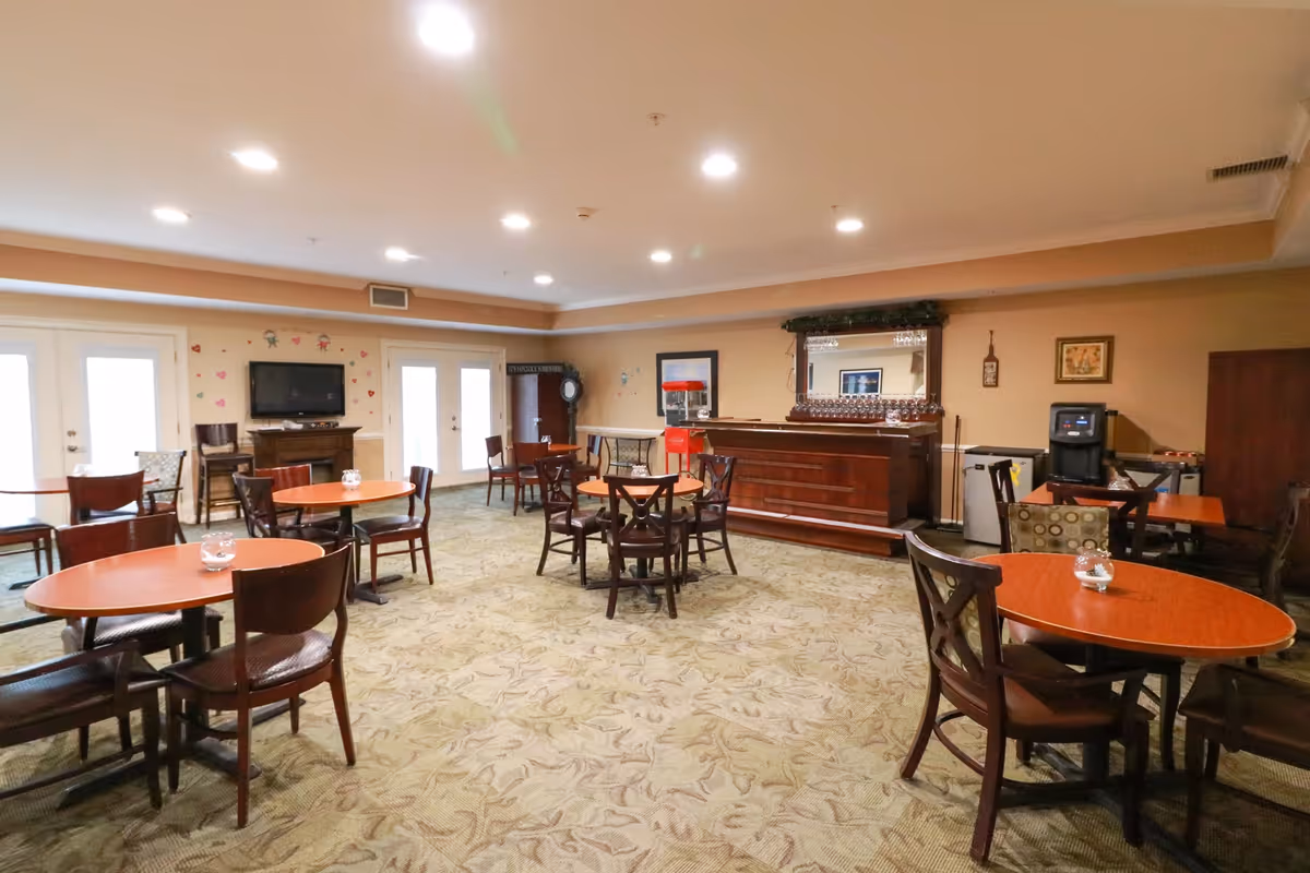 Spacious communal dining/activity room with round tables and chairs, a wooden bar, and a wall-mounted TV.