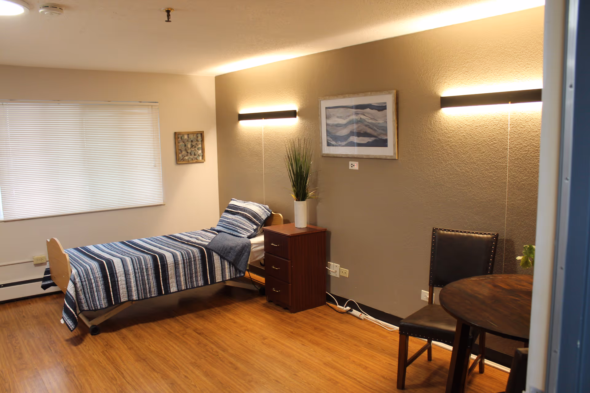A simple, clean bedroom with a single bed covered in a blue and white striped blanket. Next to the bed is a wooden nightstand with a potted plant on top. The room has wooden flooring, a round wooden table with two chairs, and two wall-mounted lights above the nightstand and table. A framed abstract artwork hangs on the wall, and a window with closed blinds is visible on the left side.