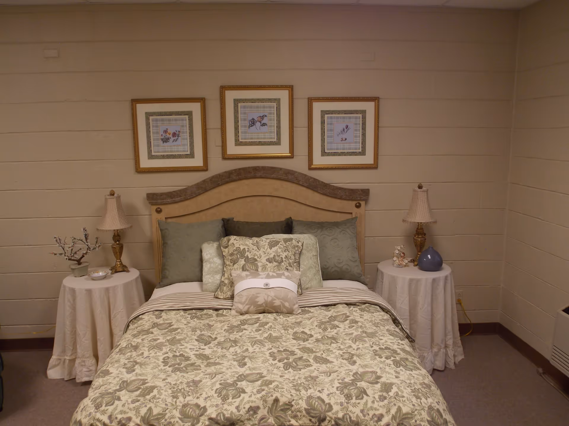 A neatly made bed with a floral patterned bedspread and multiple pillows in shades of green and beige. The bed has a curved headboard and is flanked by two round bedside tables covered with white cloths. Each table holds a decorative lamp and small decorative items. Above the bed, there are three framed floral artworks hanging on the wall. The room has beige walls and carpeted flooring.