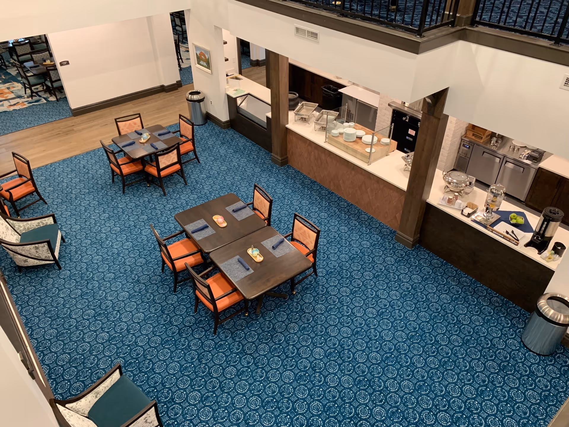 Aerial view of a dining area with several tables and orange-upholstered chairs, patterned blue carpet, and a serving counter with buffet and beverage stations.