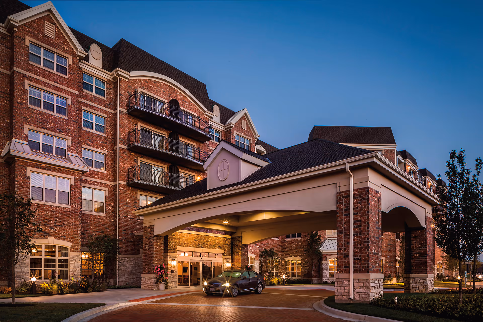 Exterior view of a multi-story brick building at dusk with a covered entrance where a car is parked. The building has multiple windows and balconies, with landscaping and trees around the entrance area.