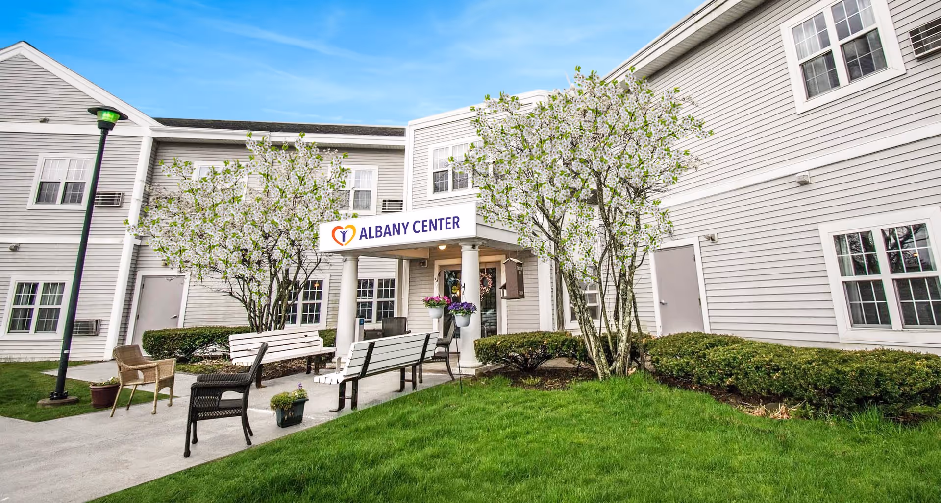 Exterior view of Albany Center Adult Home showing a two-story building with white siding, multiple windows, and a covered entrance with columns. There are blooming trees, green grass, bushes, and outdoor seating including benches and chairs along a concrete pathway.