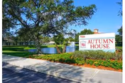 Exterior view of Autumn House facility with a large sign displaying the name 'Autumn House' near a landscaped area with green grass, bushes, and a tree. A pond and part of the building are visible in the background under a clear blue sky.