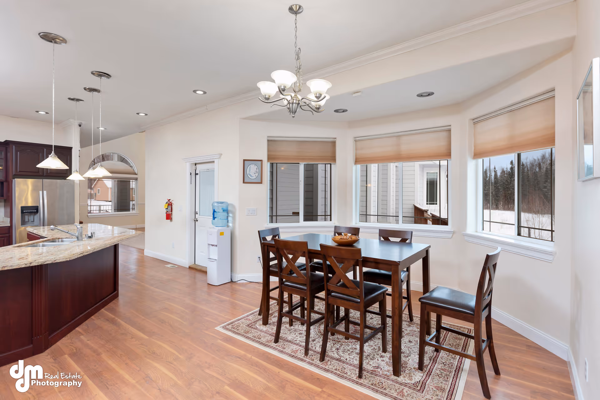 A bright dining area with a dark wooden table and six matching chairs on a patterned rug. The room has large windows with beige blinds, allowing natural light to enter. Adjacent to the dining area is a kitchen with a granite countertop island, stainless steel refrigerator, and hanging pendant lights. The floor is wooden, and there is a water dispenser and fire extinguisher near a door.